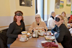 Drei Frauen sitzten an einem Tisch und haben Teller vor sich. Zwei der Frauen tragen Augenmaske.