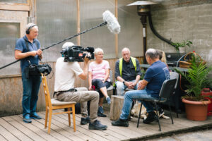 Die Gruppe sitzt um die Moderatorin herum, ein Kameramann und ein Mann mit einem Mikrofon nehmen die Scene auf.